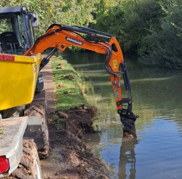 an orange digger on a canal towpath with its bucket down at an eroded, muddy bank