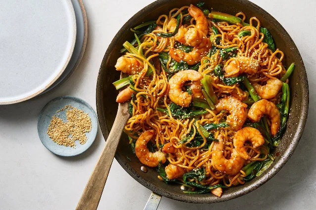 A dark skillet of noodles stained orange with chile sauce, curled pink shrimp, green stems and leaves, and scattered sesame seeds, photographed from above on a pale surface beside a small dish of sesame seeds and a stack of white plates