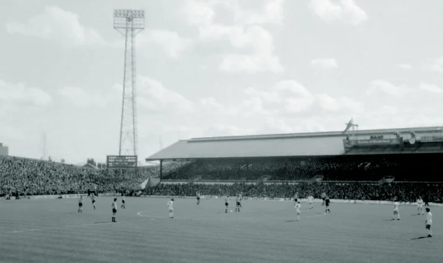"A football ground seen from the terracing, with a covered stand on the left and a crowd behind the goal"