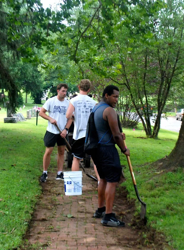 Three football players work beside a brick path with shovels and a wheelbarrow.