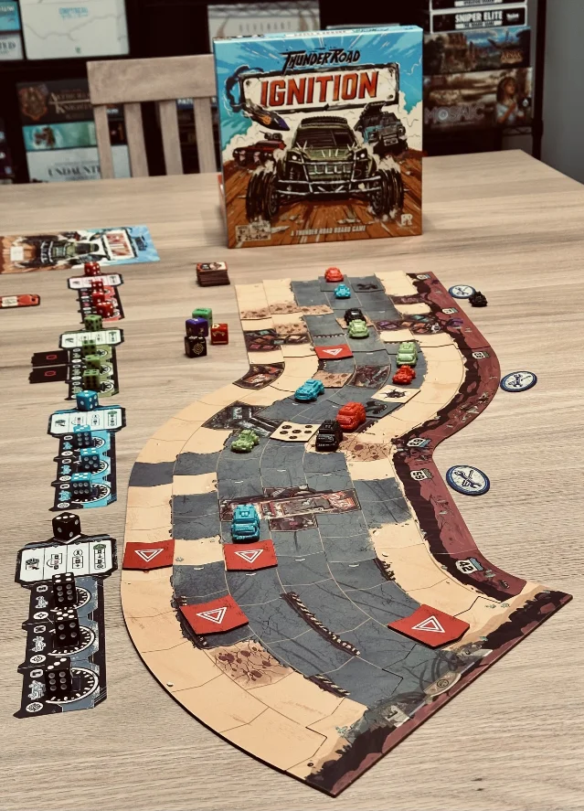 A Thunder Road: Ignition table laid out with the curved road board, miniature cars, custom dice, player dashboards and the game box