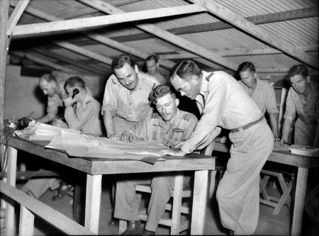 Eight men in tropical uniform around a table under a thatched roof, one holding a telephone