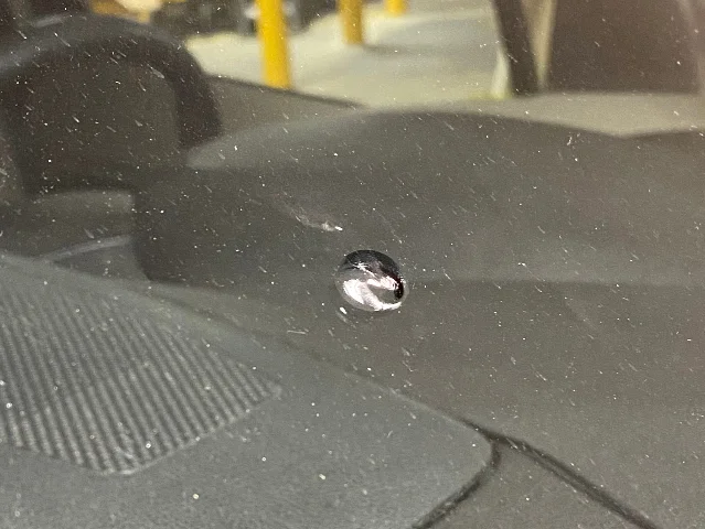 A small round rock chip in a windshield seen from outside, with a dark dashboard and a ribbed cowl panel reflected in the glass around it