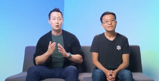 管理层合影照片：两位男士坐在浅蓝色背景前的沙发上，左侧一位穿黑色开衫正在说话，右侧一位穿黑色 T 恤、戴眼镜