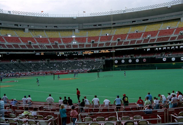 A packed baseball stadium with tiers of yellow, red and orange seats above a green artificial-turf field, players warming up on the infield