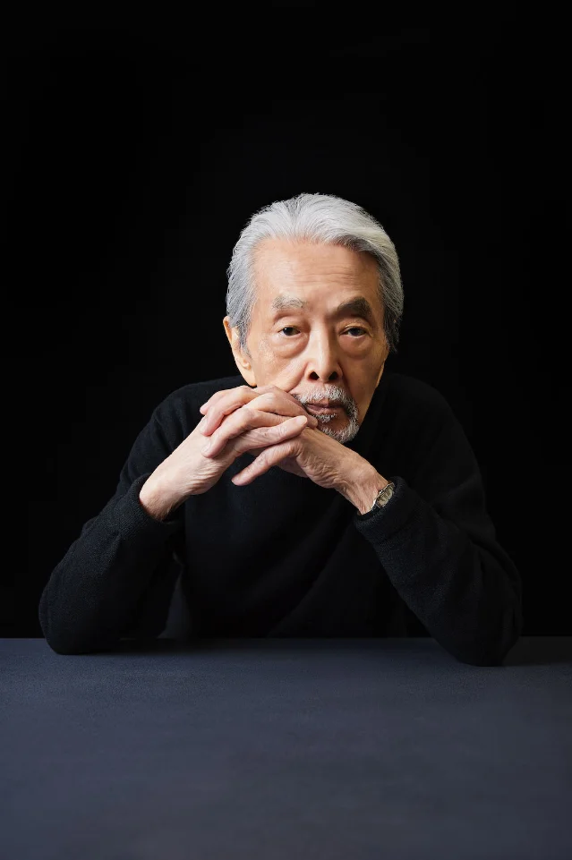 一位白发长须的老人穿着黑色毛衣，坐在深色背景前，双手交叠支在下巴前方