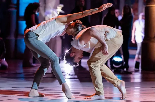 Two dancers performing on stage in casual attire, barefoot, in a lit theater space