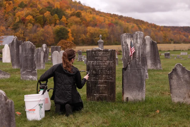 a woman kneeling in a hillside cemetery beside an old headstone, with a white bucket and a sprayer on the grass
