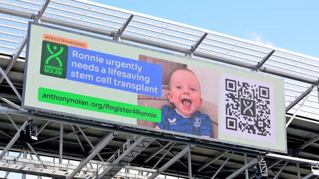 an advertising board at a football ground reading "Ronnie urgently needs a lifesaving stem cell transplant" beside a photograph of a baby and a QR code