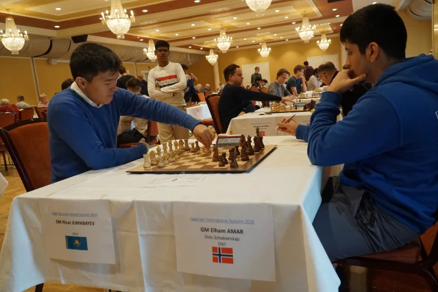 Two players at a board in a chandelier-lit hotel hall, name plates on the table in front of them