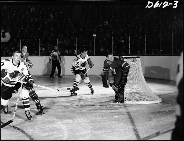 A black-and-white photograph of an indoor hockey game taken from behind the goal, with a goaltender crouched at the right of the net, three skaters in the middle distance and a crowd filling the tiered stands beyond the boards