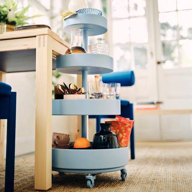 Blue IKEA PS 2026 trolley rolled partially under a dining table, holding bottles, glasses and utensils