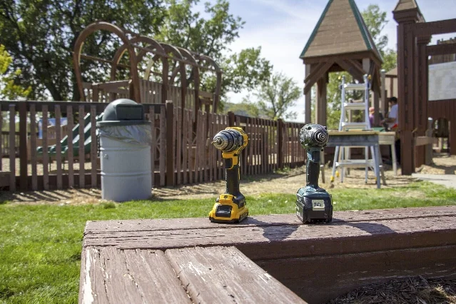 Two cordless drills standing upright on a weathered wooden rail, with playground structures and a worker on a stepladder behind them