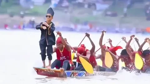 Rayyan Arkan Dikha standing at the front of a racing boat while rowers paddle behind him