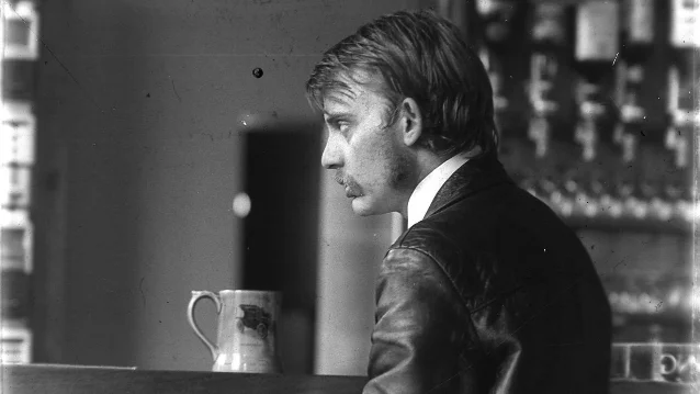 A black-and-white photograph of a young man with a moustache in a leather jacket, seen in profile at a bar counter with a ceramic mug in front of him
