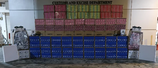 Dozens of colourful labelled boxes of vape products stacked in rows against the Customs and Excise Department wall, with loose devices and electronic components displayed on flanking tables
