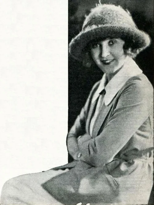 A black-and-white photograph of a smiling woman in a fur-trimmed hat and a light coat with a dark collar