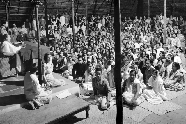A large audience of teachers seated on the ground before a raised platform