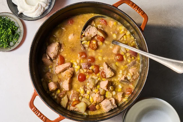 A Dutch oven filled with salmon, corn, tomatoes and potatoes, with chopped herbs and dairy on the side