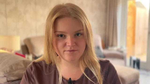 a young woman with long blonde hair sitting indoors, looking towards the camera