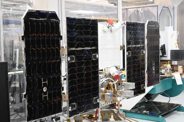 A small satellite bus on a bench in a clean room with four dark solar panel wings unfolded out to its sides