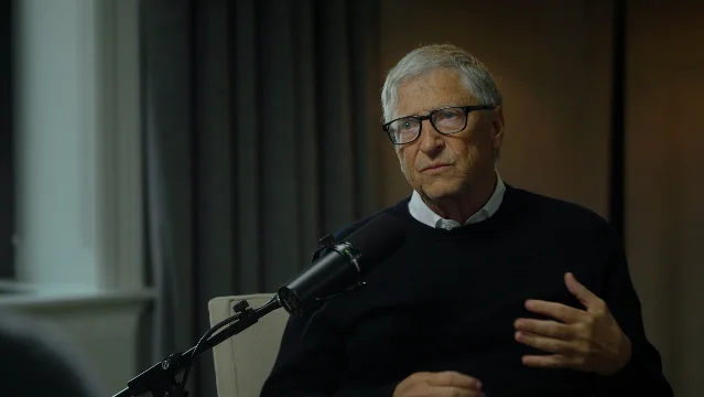 比尔·盖茨在《The Ezra Klein Show》录制现场,手持麦克风,穿深色毛衣与浅色衬衫