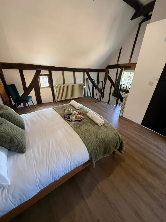 A bedroom under a sloping ceiling, with a double bed with white linen and a green throw, a tea tray, a green armchair and exposed dark timber beams above a stairwell