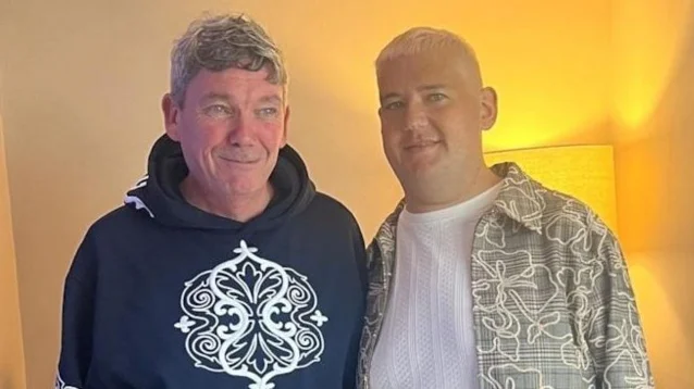 Two men standing close together indoors, one in a dark hoodie and one in a patterned shirt