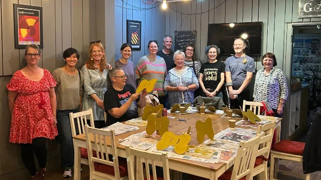 A group of volunteers standing around a wooden table covered with newspaper, paint pots and freshly painted gold bows