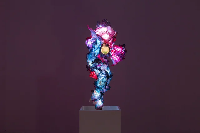 A brightly lit multicoloured sculpture made from crumpled blue, pink and white algae-based material on a pedestal