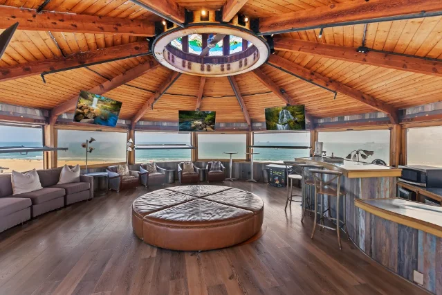 A round living room under redwood beams, with a stained-glass cupola at the centre of the ceiling, sofas set against wide windows, and the Pacific beyond them