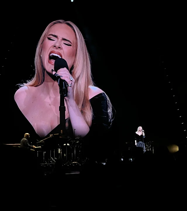 Adele singing on stage, shown large on an arena screen with the audience in front of her