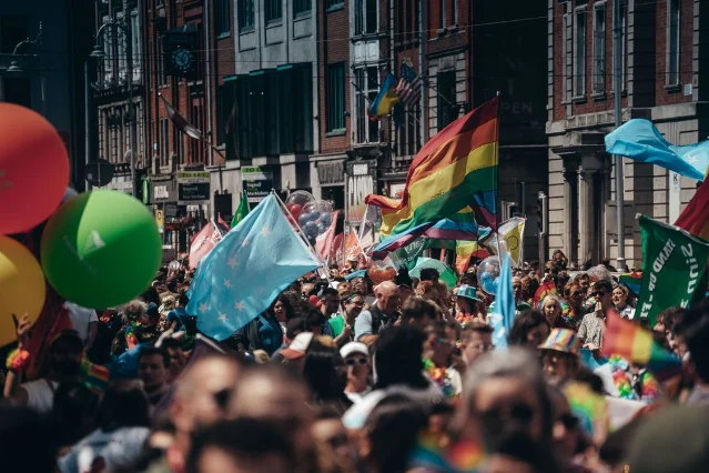 A colorful Pride parade crowd waves rainbow flags and balloons on a sunny city street