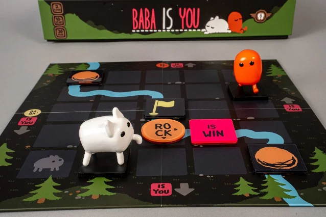 Baba Is You tabletop physical setup with rule tiles, figurines, and grid board