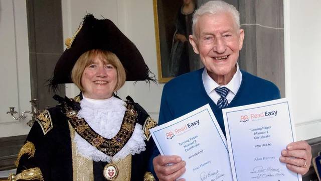 an elderly man in a blue sweater holding two printed certificates beside a woman in dark ceremonial robes and a chain of office