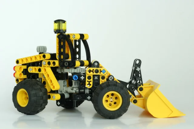 LEGO Technic shovel loader model in yellow and black, showing exposed beam-and-connector mechanical assembly on a light grey background