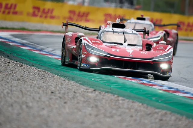 A red Toyota TR010 prototype racing through a corner with its headlights on, a second red prototype behind it and a DHL barrier in the background