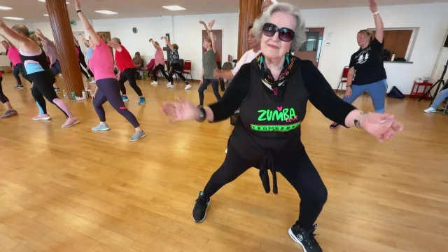Eighty-two-year-old Necla wears sunglasses and a black Team Berna top while leading dancers on a wooden hall floor.