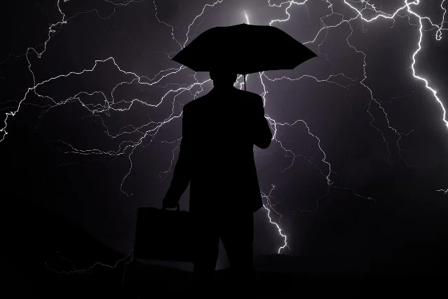 经济衰退概念图:暴风雨中撑伞的商人剪影,背景是下行的图表走势
