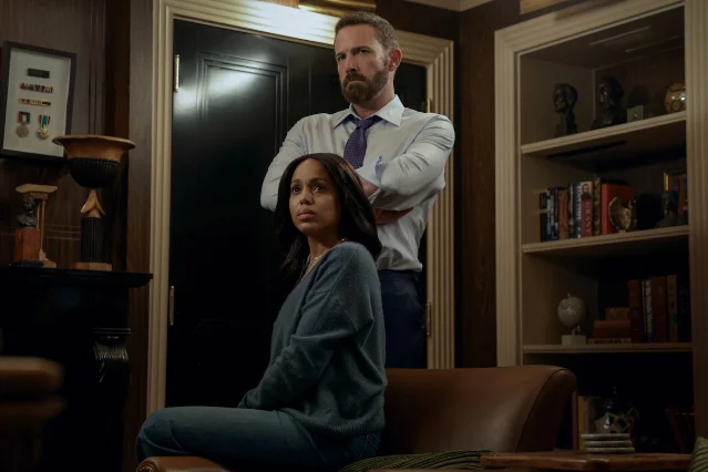 A woman seated in a chair in a wood-panelled study while a man stands behind her with his arms crossed