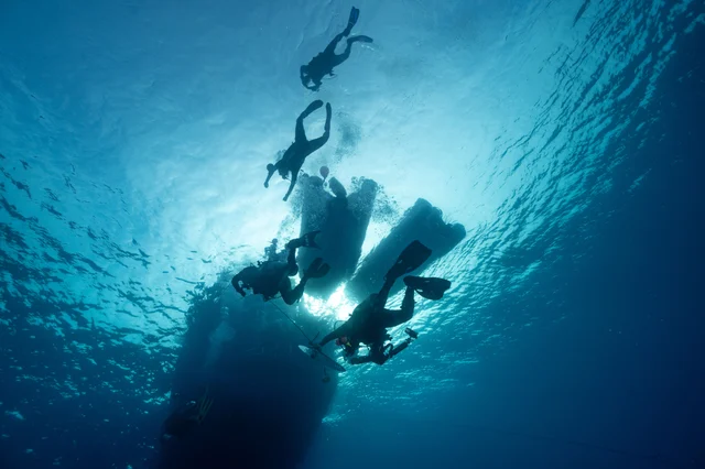 Silhouetted scuba divers swimming underwater beneath a yacht in clear blue water