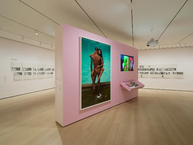 展厅里韦克斯的黑白照片面板分挂两面白墙，中间隔着一面粉色墙，粉色墙右侧挂着一本摊开的杂志