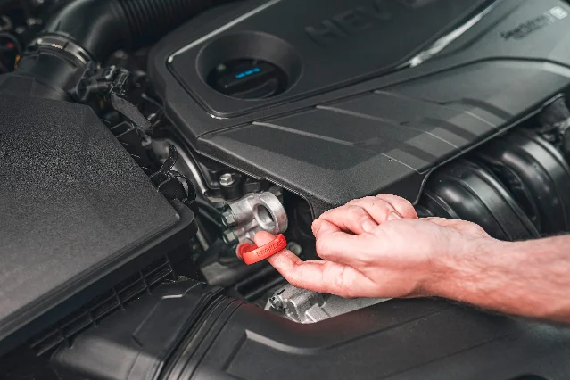 A hand holding a red filler cap stamped with the words ENGINE OIL beside the engine cover of an open engine bay