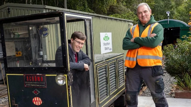 Two men smiling beside a small black locomotive carrying a Woodhorn Station sign, one seated in the cab and one standing on the platform in a hi-vis vest