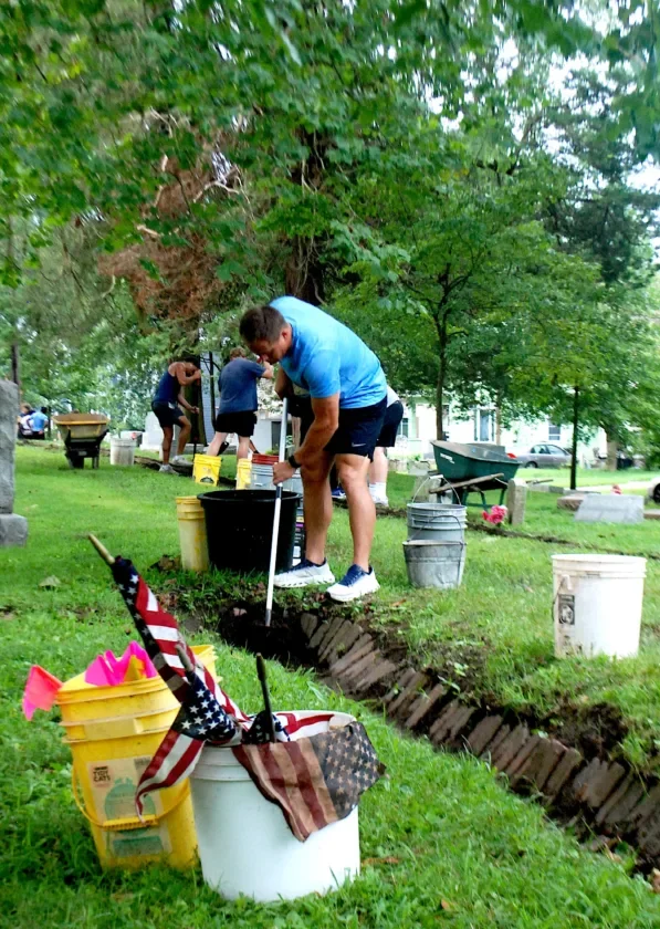 A man in a light blue shirt clears a drainage channel beside cemetery markers.