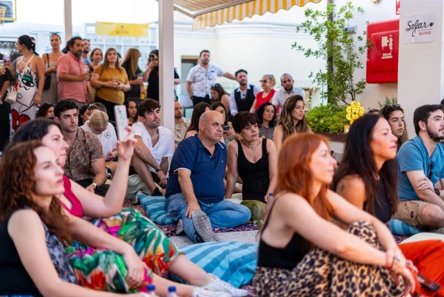 A crowd lounges on a sun-dappled lawn at an outdoor free concert, trees overhead, stage in the distance