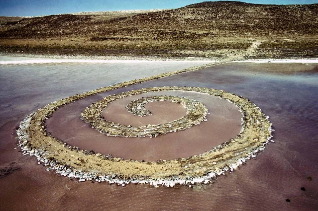 黑色石块构成的《Spiral Jetty》盘入褐红色湖水，远处是裸露山坡。