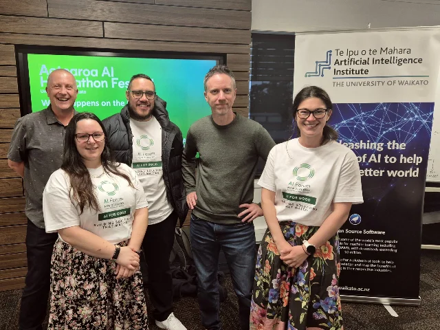 Five Wise Group team members stand beside Aotearoa AI Hackathon Festival and University of Waikato signage