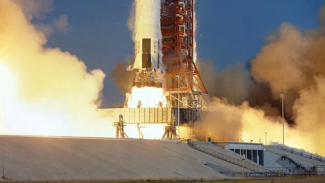 A Saturn V lifting off from its pad, its engines firing and the launch structure beside it