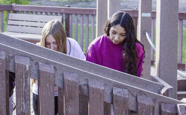 Two teenagers leaning over a weathered wooden railing, one in a white top and one in a magenta sweatshirt